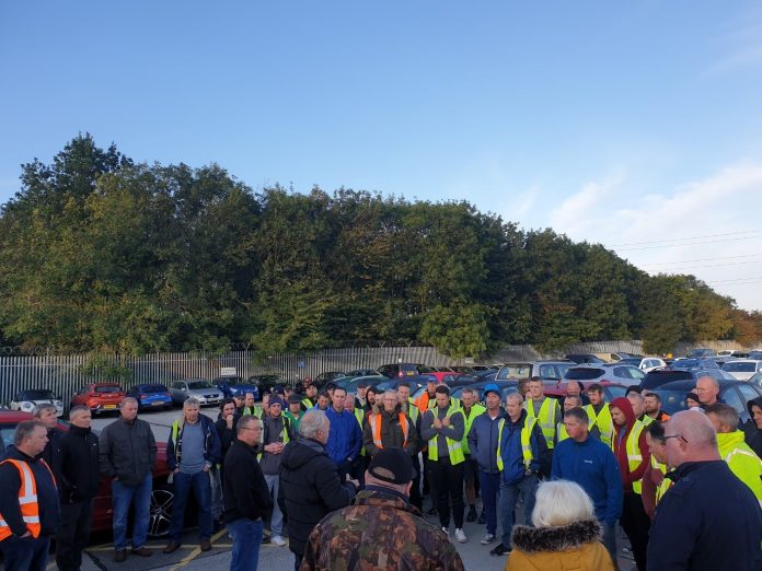 striking workers, wearing work clothes, stand gathered in a workplace car park