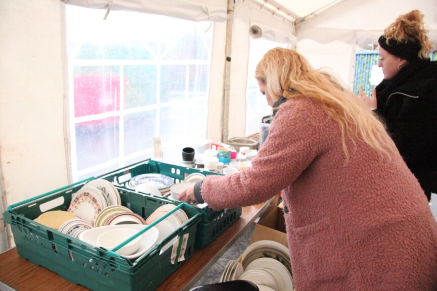 Someone with long blonde hair and a pink coat browses through a green crate full of crockery