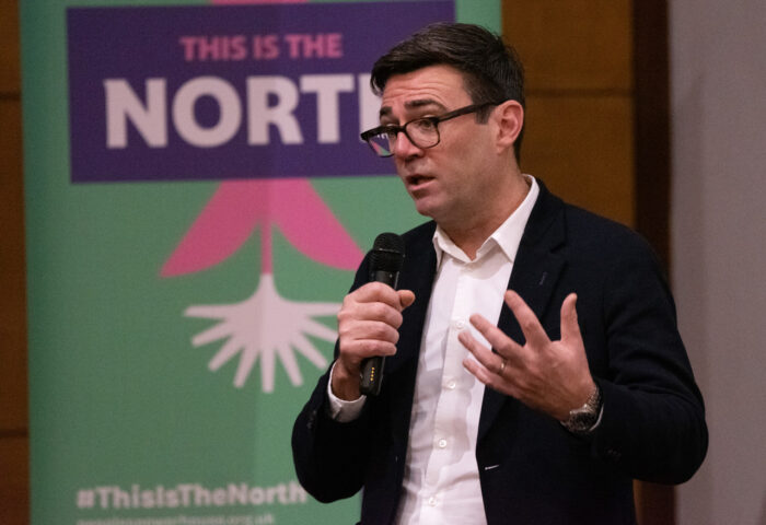 Andy Burnham is on stage at this is the North conference, addressing the crowd using a persuasive gesture. He wears black-rimmed glasses, a dark suit jacket and white shirt (no tie).