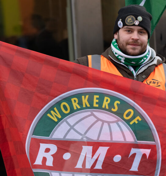 A railway worker wearing winter clothes and a hi-vis vest holds up a red 'workers of RMT' flag