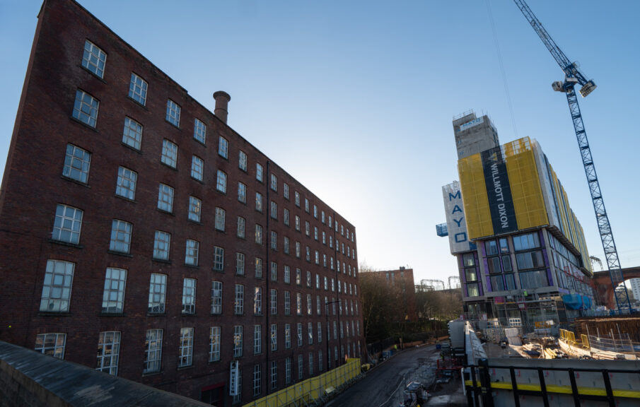 Stockport Hat Museum on the left of the shot, in an imposing mill building, next to the bus station. The sky is clear and blue, and the sun is just behind the mill.