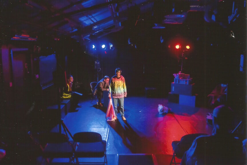 Zoe is performing on stage in a scene with dramatic lighting. She's wearing a casual outfit and looking at the camera while others in the scene's background look on.