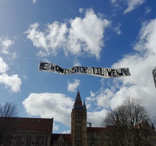 A banner reading "we won't stop 'til we win" flies over the Whitworth building at the University of Manchester. It's a neo-gothic building with a bright blue sky with fluffy white clouds above it.