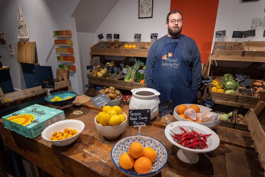 Ashley, a white man with glasses and a beard, wearing a dark blue apron with his company's logo, stands inside his greengrocer shop with trays and bowls of fruit & veg on display.