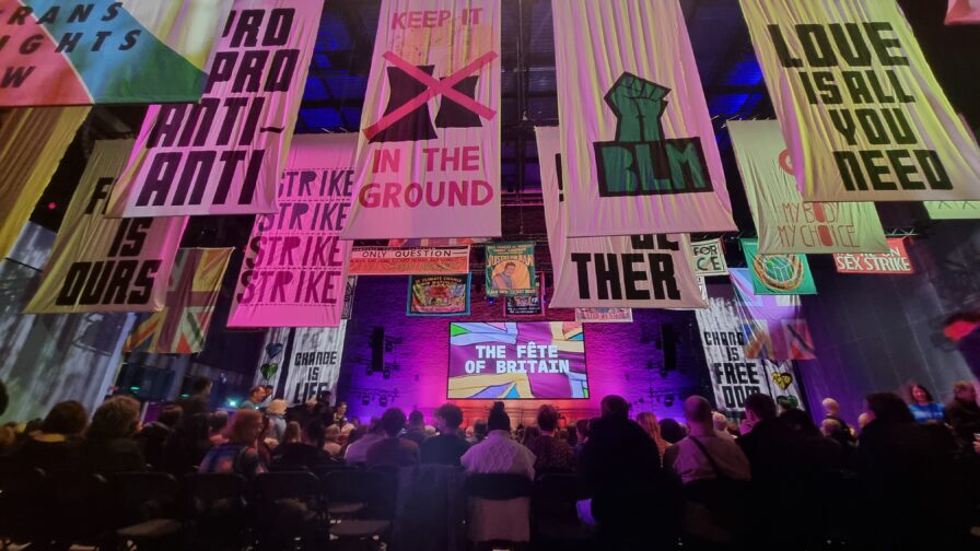 A view of the stage at Factory exhibition centre, taken from the rear of the cowd and looking up at banners with activist messages. The room is dimly-lit in a purple hue.