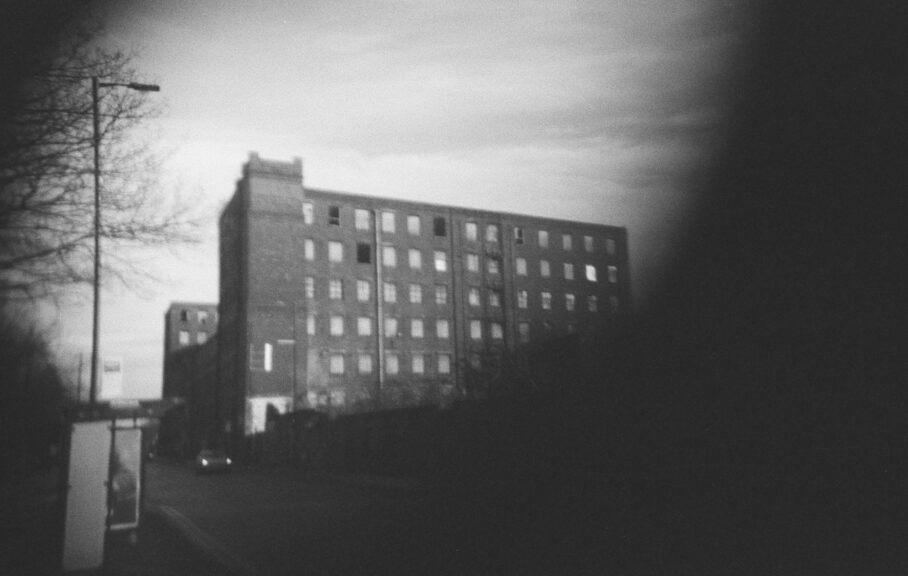 A black-and white photo of Brunswick Mill, a 7-storey Victorian mill with internal courtyard. To the left foreground is a bus stop, a lamppost, and overhanging branches from the trees on the adjoining plot just out of sight.