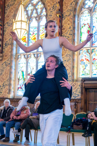 One performer sits on another's shoulders as part of the Zugzwang dance performance. The audience can be seen in the background, as well as the spectacular and detailed decor in the room - there are arched stained glass windows and gold-coloured floral patterns all over the walls.