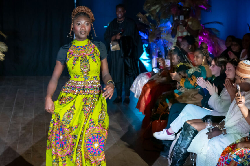 A black woman walks in a fashion show, wearing a yellow dress with traditional patterns in blue, pink and gold. She wears bangles on her left hand, large triangular earrings, and light brown braids coiled on top of her head.