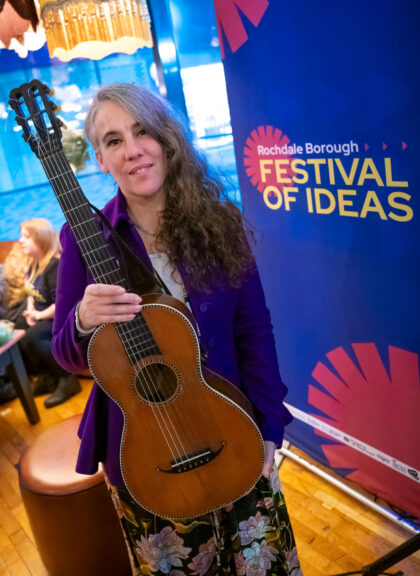 A white person in a blue cardigan with long, greying brown hair, carrying a stringed musical instrument, stands next to a large banner for Rochdale Festival of Ideas