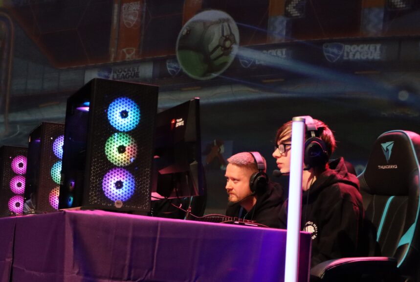 Two contestants wearing headphones sit at a table with a purple cloth, looking at a computer screen. There are speakers with RGB colour-change lighting at the front of the table, and arena banners in the background for 'Rocket League'.