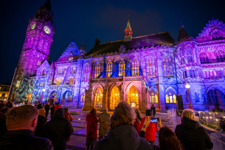 Rochdale Town Hall, a neo-gothic building with a clock tower, is lit up at night in blues, purples and orange.