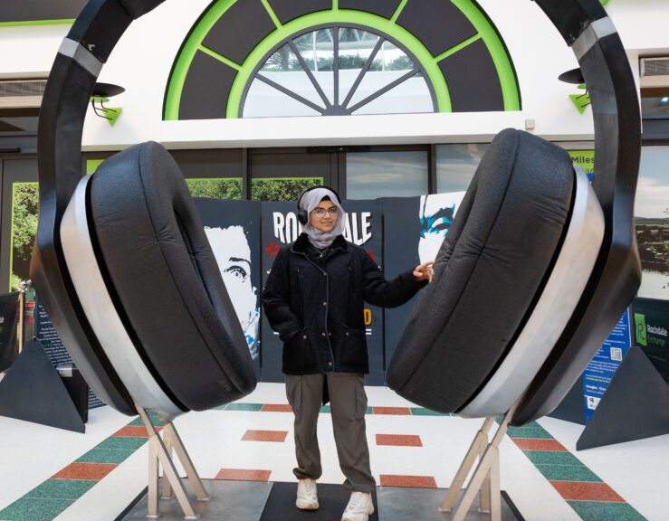 A young person wearing outdoor clothing and a hijab stands within a giant pair of headphones in front of posters for the Rock Dale Wall of Sound