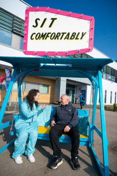 Two people sit having a conversation on a blue swing chair set up outdoors. The swing chair structure has a big white sign with a pink border on top, it reads "sit comfortably".