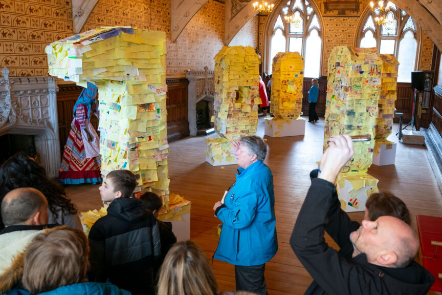 Adults and children walk around a room in Rochdale Town Hall displaying children's ideas for a future Rochdale on Post-Its stuck to columns set up throughout the space.