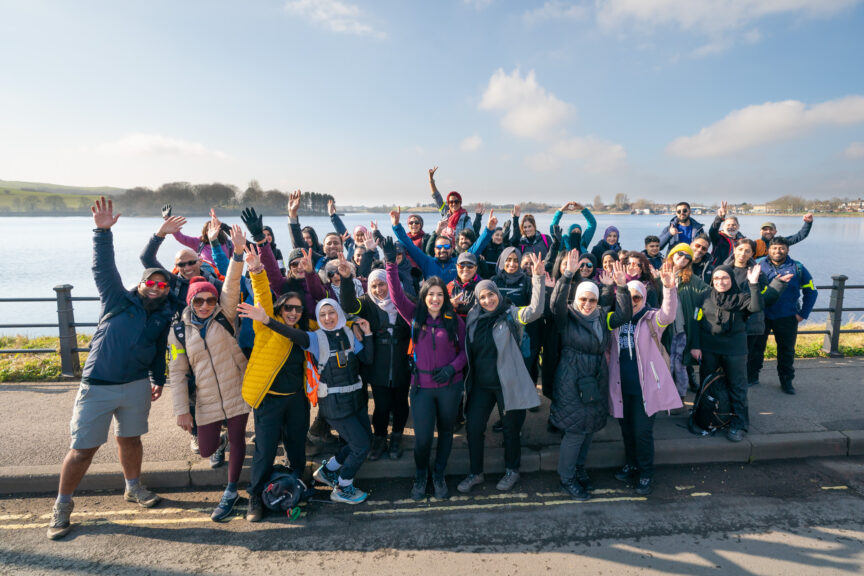 About 40 people in outdoor clothing pose for a photo in front of a body of water with trees, green hills, and some houses on the horizon. It's sunny and there is a blue sky with fluffy white clouds.