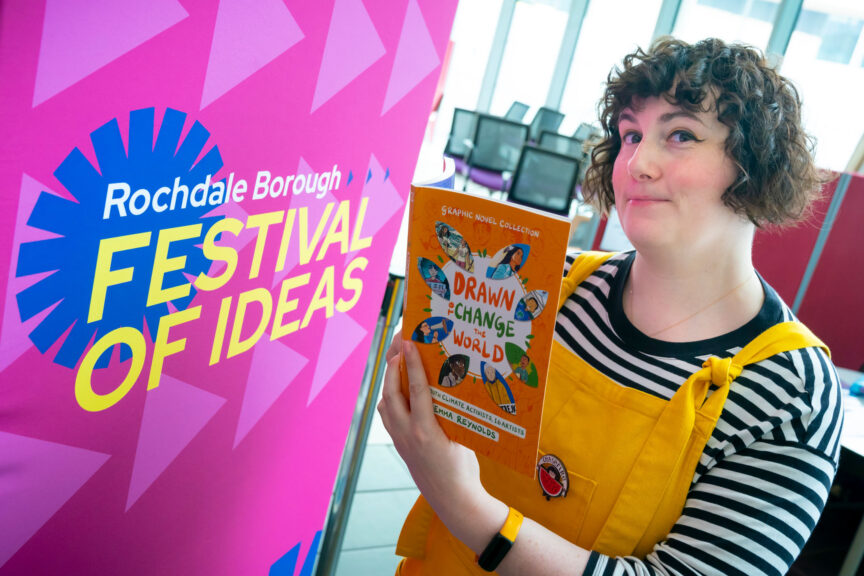 A white person with chin-length dark curly hair, wearing a black and white striped t-shirt and yellow dungarees, stands next to a sign for the Festival of Ideas.