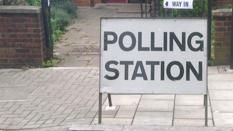 A freestanding sign reading 'POLLING STATION' in black text on a white background, on a paved surface with a small part of a builing and some foliage visible in the background.