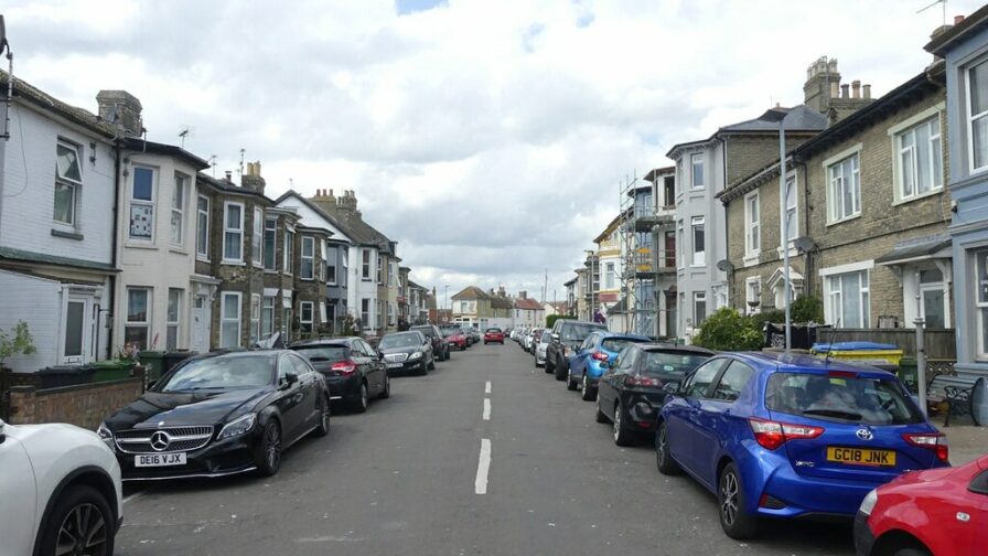 A street of Victorain terraced housing in varying styles, with houses and cars lining both sides of the street. One house has scaffolding on it, and the sky is blue with lots of clouds.