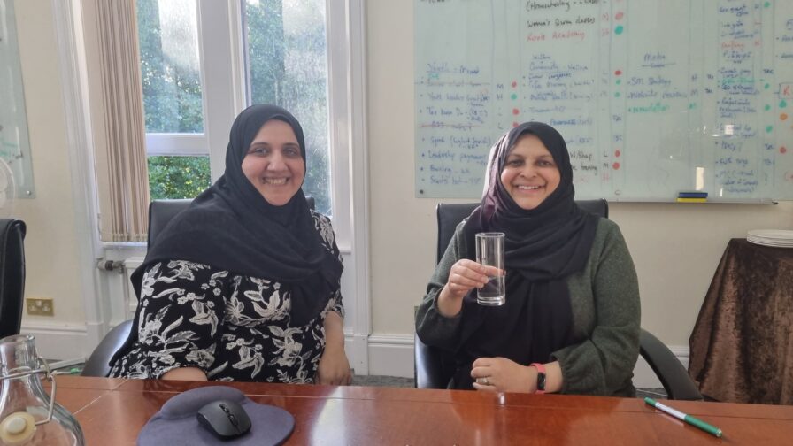 Halima and Robina from the British Muslim Heritage Centre. They are two young, smiling hijabi women sat at a table in front of a large window in a light-coloured room.