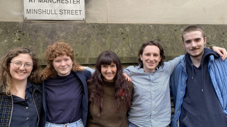 The Manchester Five posing for a photo in front of a wall with a metal sign engraved with "The Crown Court at Manchester Minshull Street". They are a group of white people dressed in smart/casual clothing. Their arms are around each other's shoulders and they are all smiling.