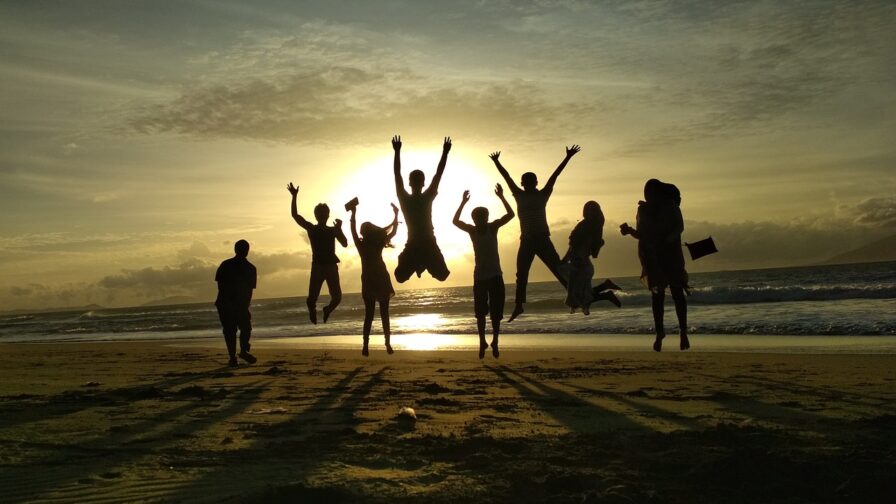 A group of people on a sandy beach with the tide out, jumping into the air. The sun is setting, and the people can only be seen in silhouette.