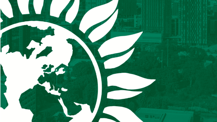 The Green Party logo (the earth surrounded by petals as if it were the centre of a sunflower) on a green background with a transparent overlay of the Manchester skyline.