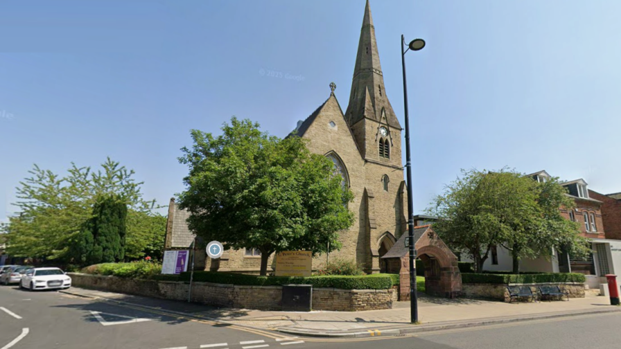 A sandstone church with a spire and trees in the churchyard, on a main road through Manchester, UK. It is a bright, sunny day with a clear, blue sky.